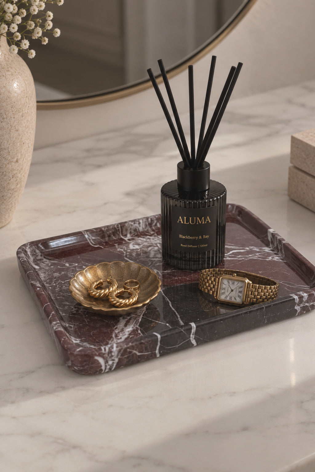 A reed diffuser bottle labeled ALUMA with black reeds, displayed on a dark marble tray alongside gold jewelry and a watch on a light marble countertop.