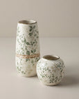 Two ceramic vases with a delicate green floral pattern on a warm off-white background, one tall and one short, set against a neutral beige studio backdrop.