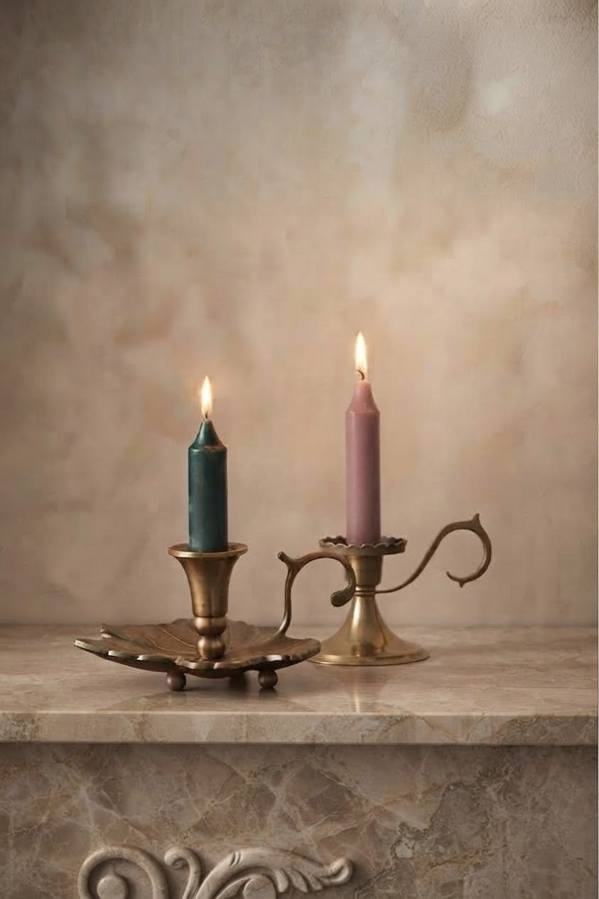 Two lit taper candles in ornate brass candlesticks, one teal and one dusty pink, displayed on a stone mantel against a textured beige plaster wall.