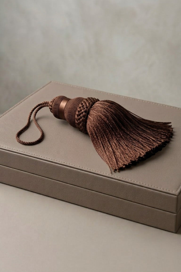 A brown braided tassel with a matching cord lies on top of a taupe-colored rectangular box against a neutral textured background.