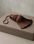 A brown braided tassel with a matching cord lies on top of a taupe-colored rectangular box against a neutral textured background.