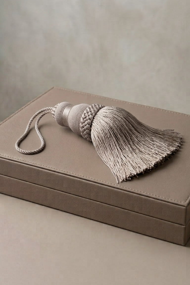 A large decorative tassel with a braided top and long fringe, resting on a taupe-colored box against a neutral beige background.