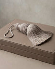 A large decorative tassel with a braided top and long fringe, resting on a taupe-colored box against a neutral beige background.