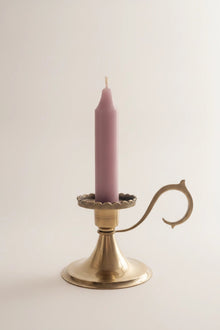 A single lit taper candle in a dusty mauve-pink color, set in a polished brass candlestick holder with a decorative curved arm, against a plain off-white background.