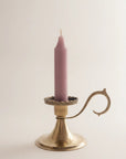 A single lit taper candle in a dusty mauve-pink color, set in a polished brass candlestick holder with a decorative curved arm, against a plain off-white background.