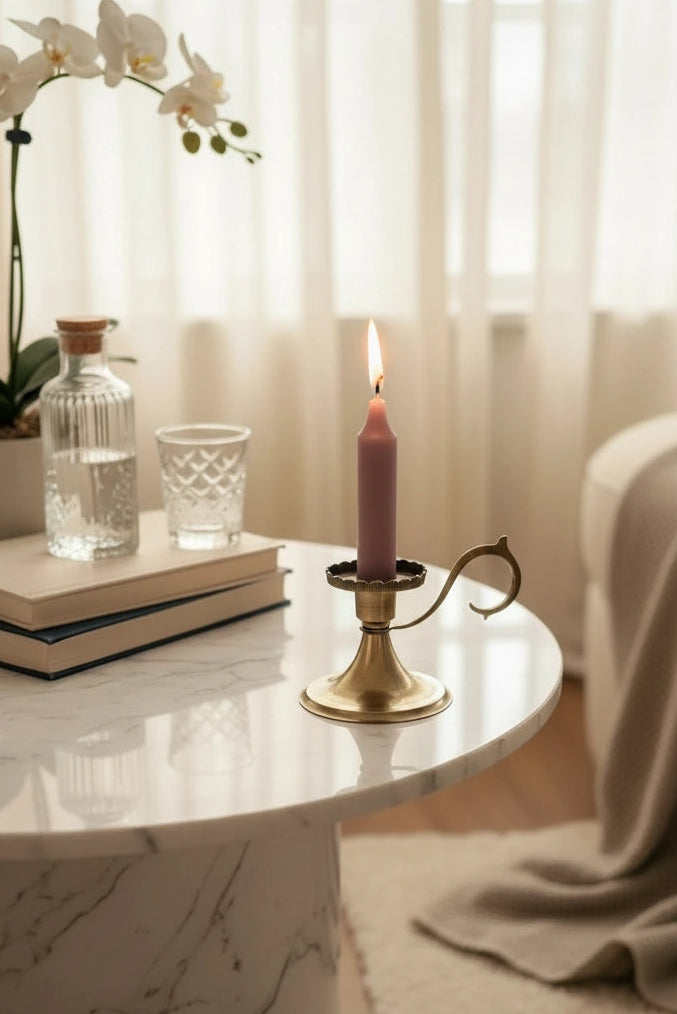 A lit pink candle in a gold-toned metal holder sits on a white marble side table in a softly lit bedroom. A glass bottle, small glass, books, orchid flowers, sheer curtains, and a draped beige blanket are visible in the background.