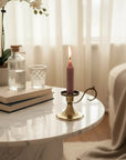 A lit pink candle in a gold-toned metal holder sits on a white marble side table in a softly lit bedroom. A glass bottle, small glass, books, orchid flowers, sheer curtains, and a draped beige blanket are visible in the background.