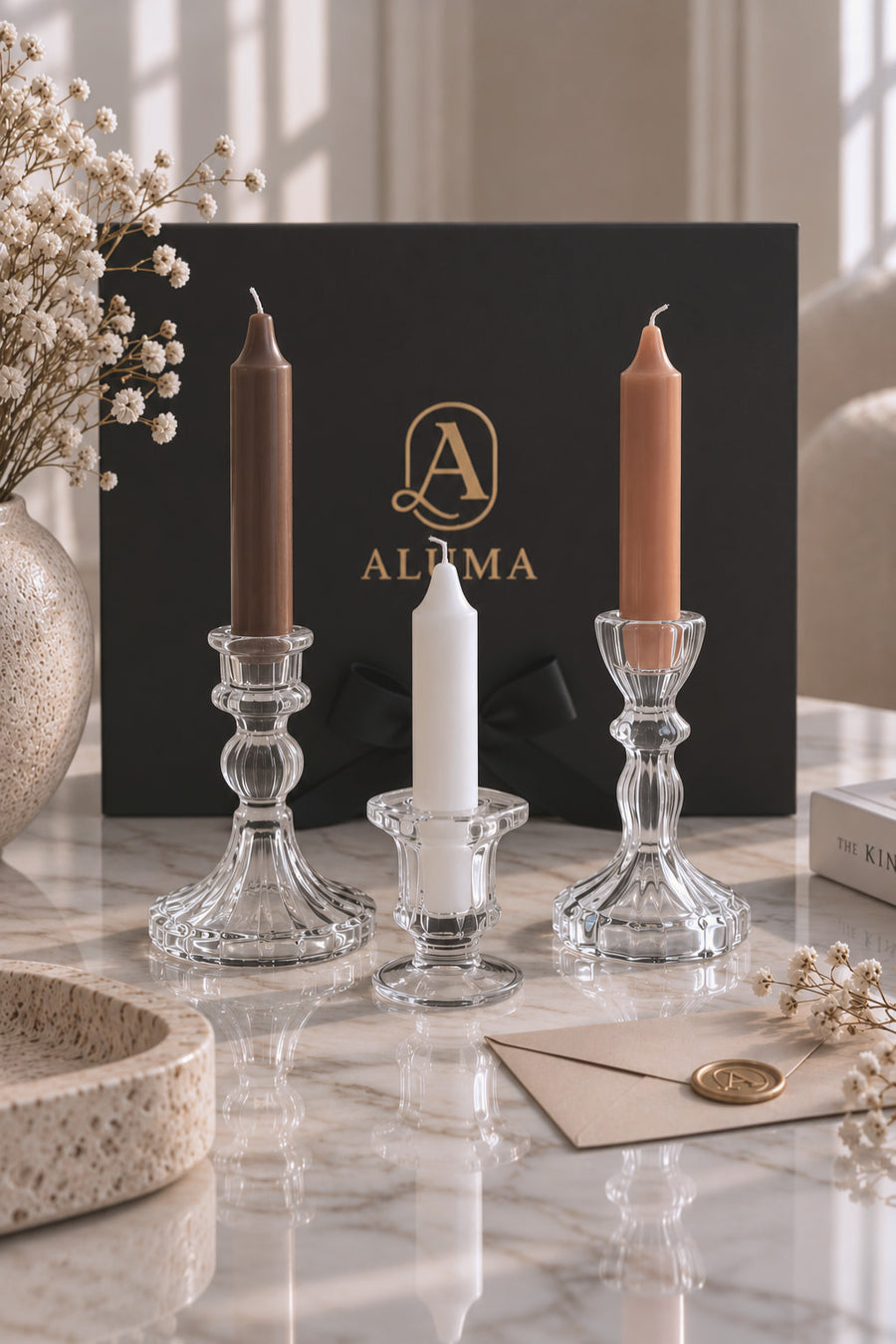 Three glass candlesticks holding taper candles in brown, white, and orange tones, arranged on a marble surface in front of a black ALUMA box with a gold logo.