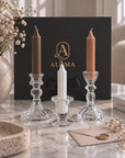 Three glass candlesticks holding taper candles in brown, white, and orange tones, arranged on a marble surface in front of a black ALUMA box with a gold logo.