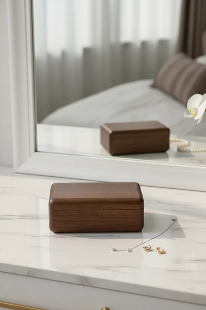A rectangular dark brown wooden jewelry box with a hinged lid, resting on a white marble surface in front of a large white-framed mirror. A reflection of the box is visible in the mirror, along with a pearl necklace and small gold earrings on the surface.