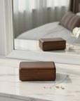 A rectangular dark brown wooden jewelry box with a hinged lid, resting on a white marble surface in front of a large white-framed mirror. A reflection of the box is visible in the mirror, along with a pearl necklace and small gold earrings on the surface.