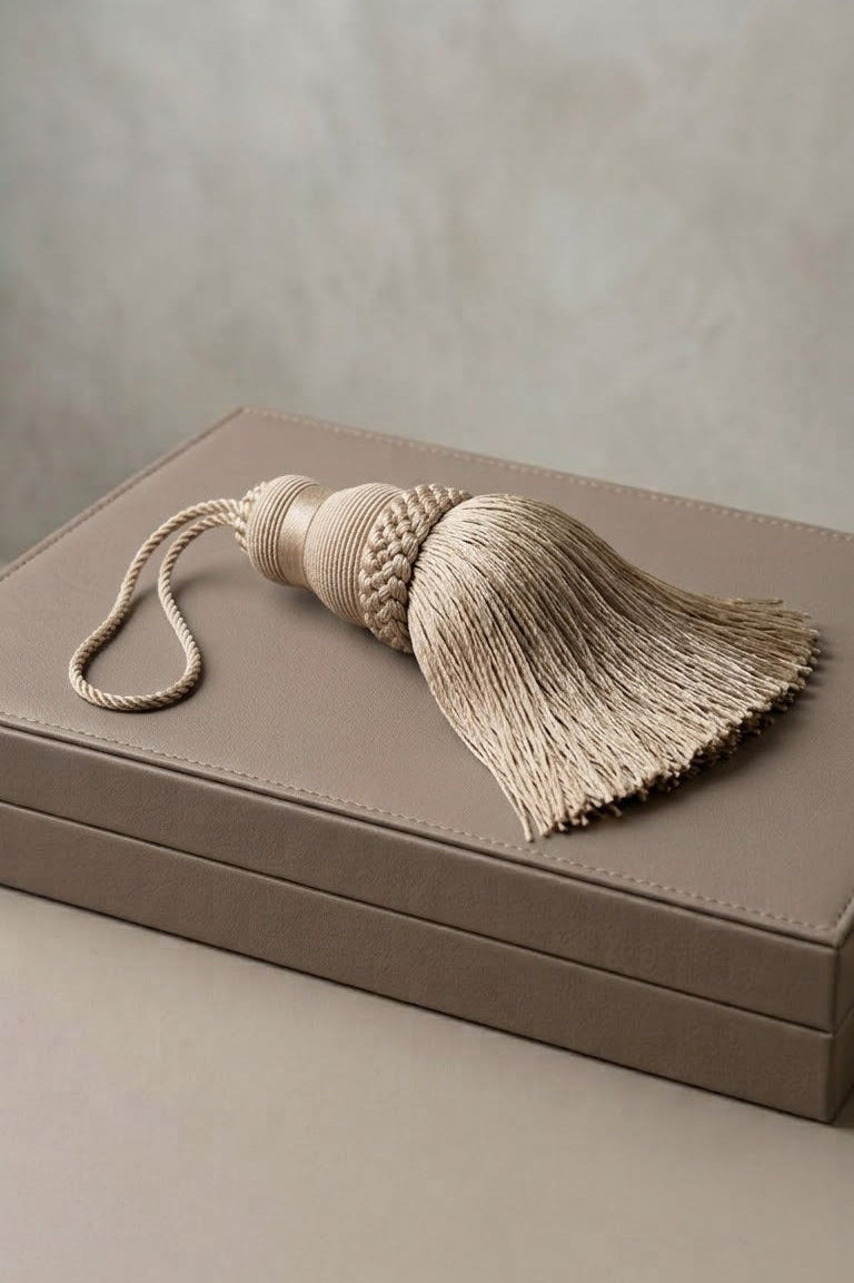 A beige decorative curtain tassel with a braided top and loop, resting on a matching taupe leather box against a neutral textured background.