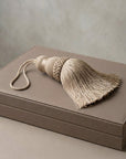 A beige decorative curtain tassel with a braided top and loop, resting on a matching taupe leather box against a neutral textured background.