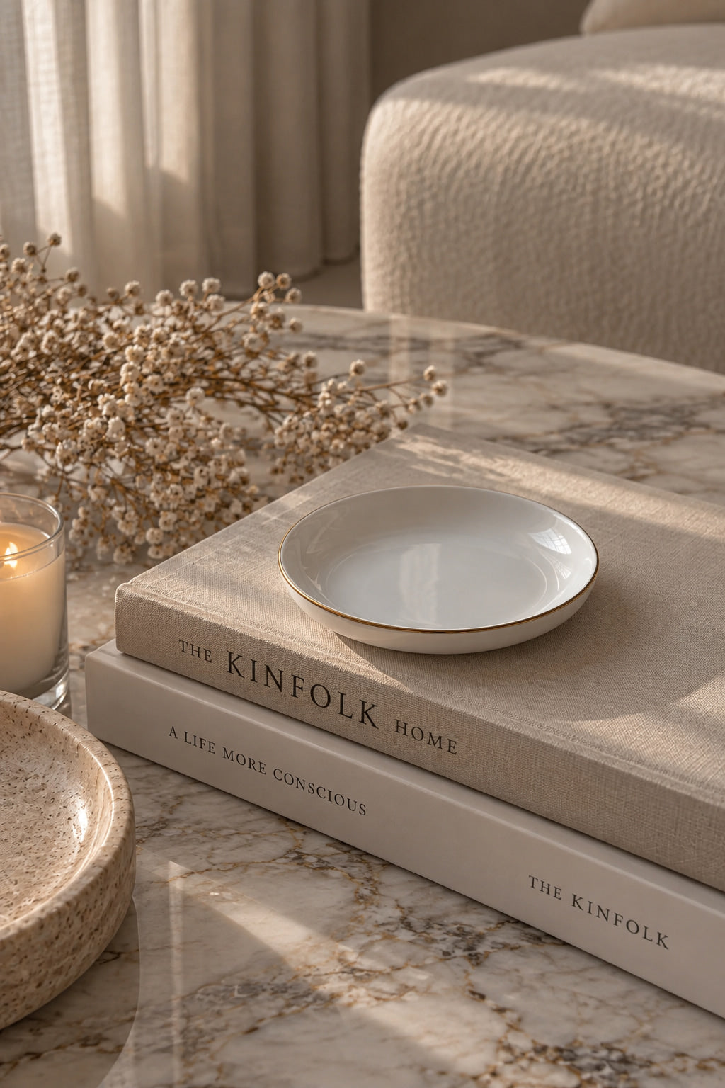 A small round white ceramic dish with a delicate gold rim sits on top of two stacked books on a marble table, styled with dried flowers, a candle, and another dish in a softly lit home interior.