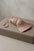 A dusty pink and beige embroidered tassel with a matching cord lies on top of a taupe leather jewelry box against a neutral textured background.