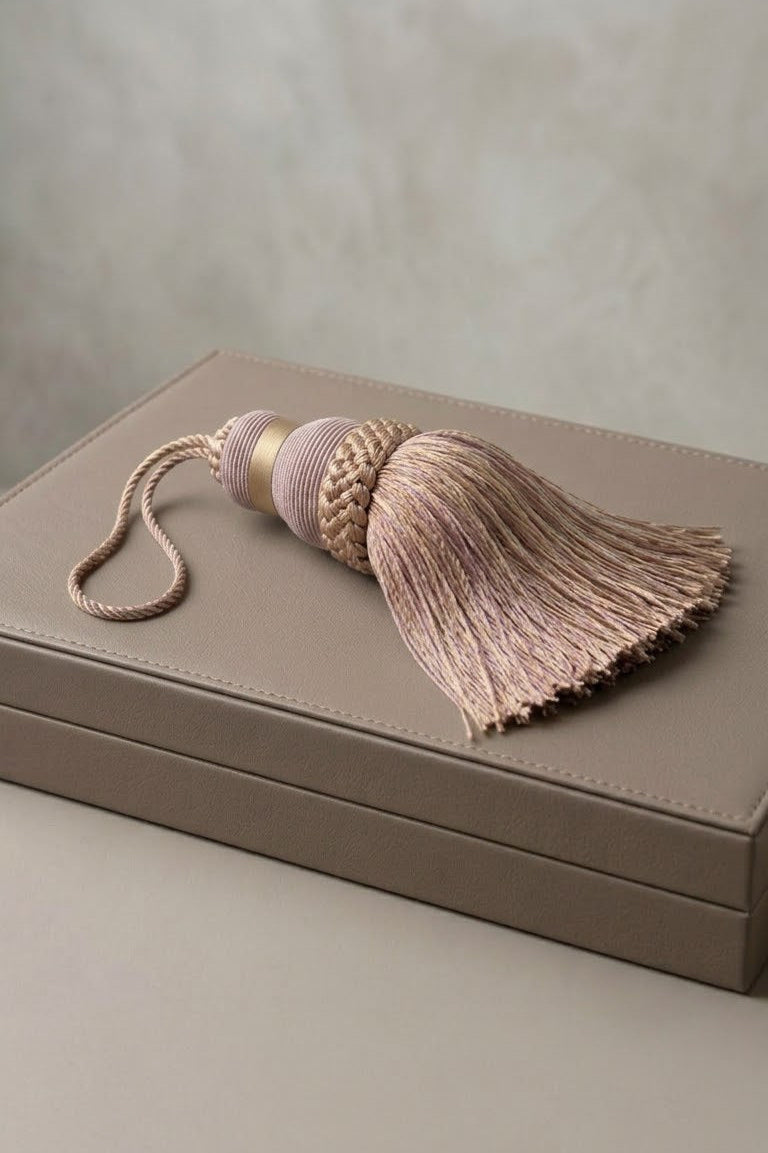 A dusty pink and beige embroidered tassel with a matching cord lies on top of a taupe leather jewelry box against a neutral textured background.