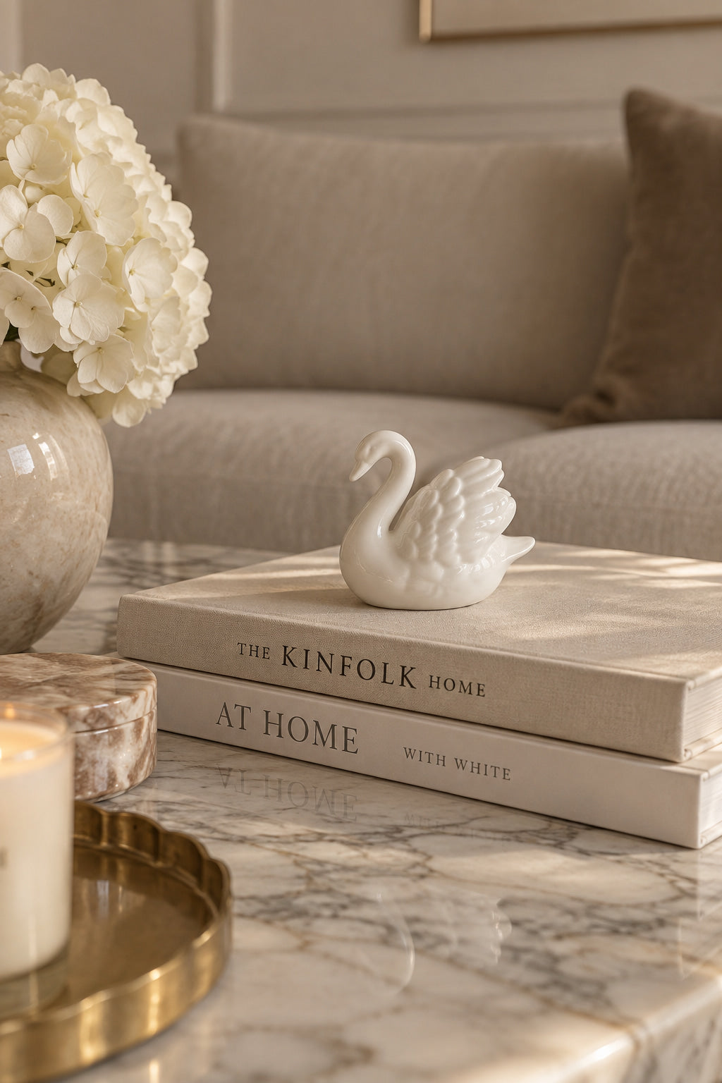 ceramic white swan  on top of tow home decor books, on top of a white marble  table, next to flower vase, cream background  and cream sofa at the conner.