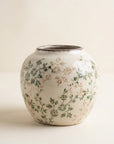 A small ceramic vase with a rounded body and short flared rim, decorated with a delicate hand-painted floral pattern in green and muted tan on an off-white glaze, set against a plain cream background.