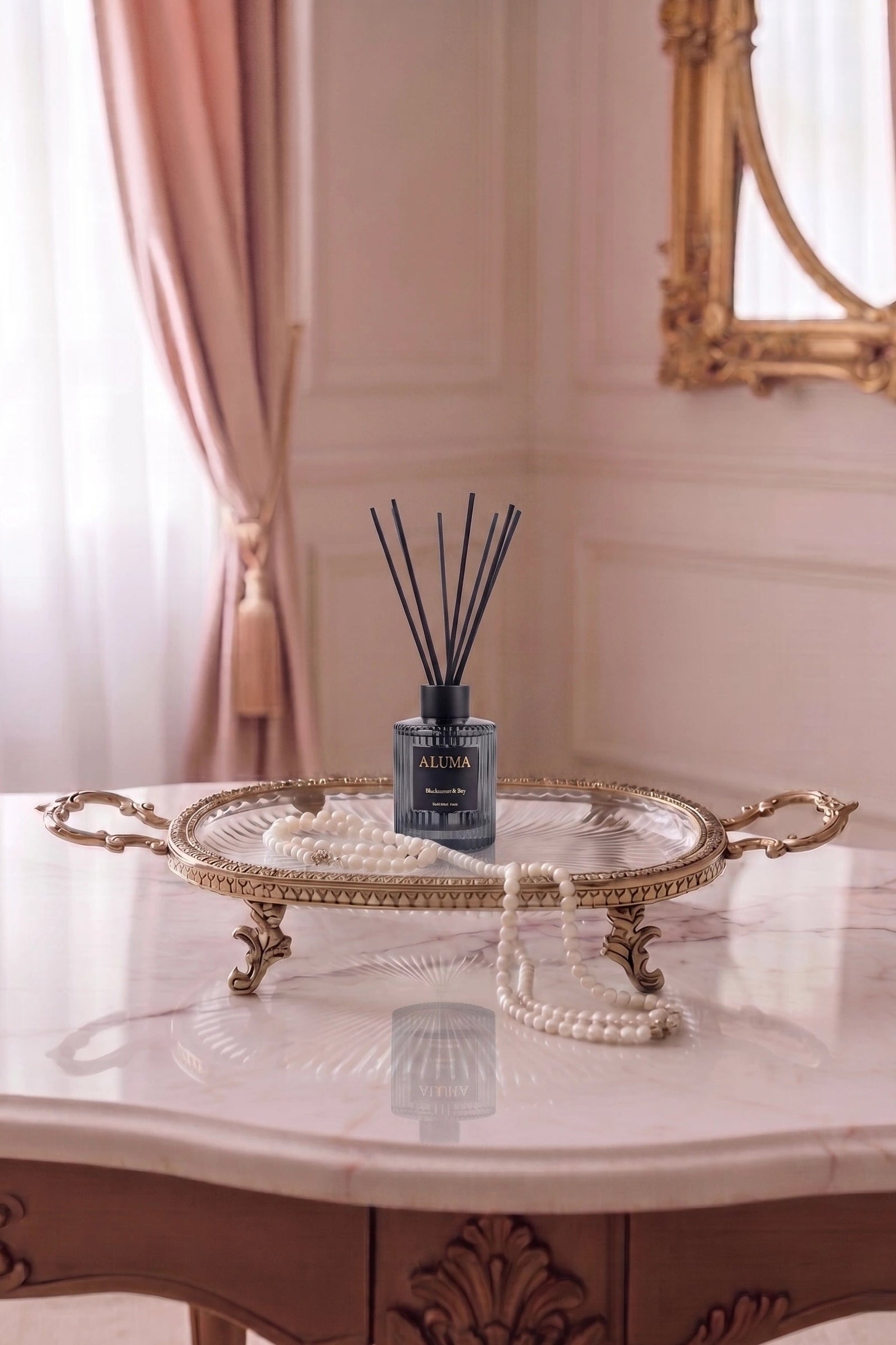 A reed diffuser bottle with black reeds sits on an ornate gold tray draped with a pearl necklace, placed on a marble table in a softly lit room with pink curtains and a gold mirror.