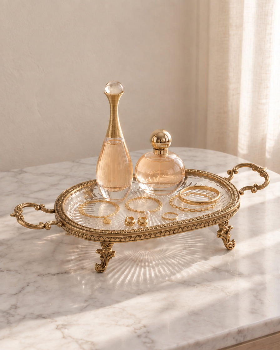 An ornate gold metal tray with decorative handles and legs sits on a white marble table, holding two clear glass perfume bottles and several gold jewelry pieces. The scene is softly lit by natural light from a nearby window, with a neutral beige wall and curtain in the background.