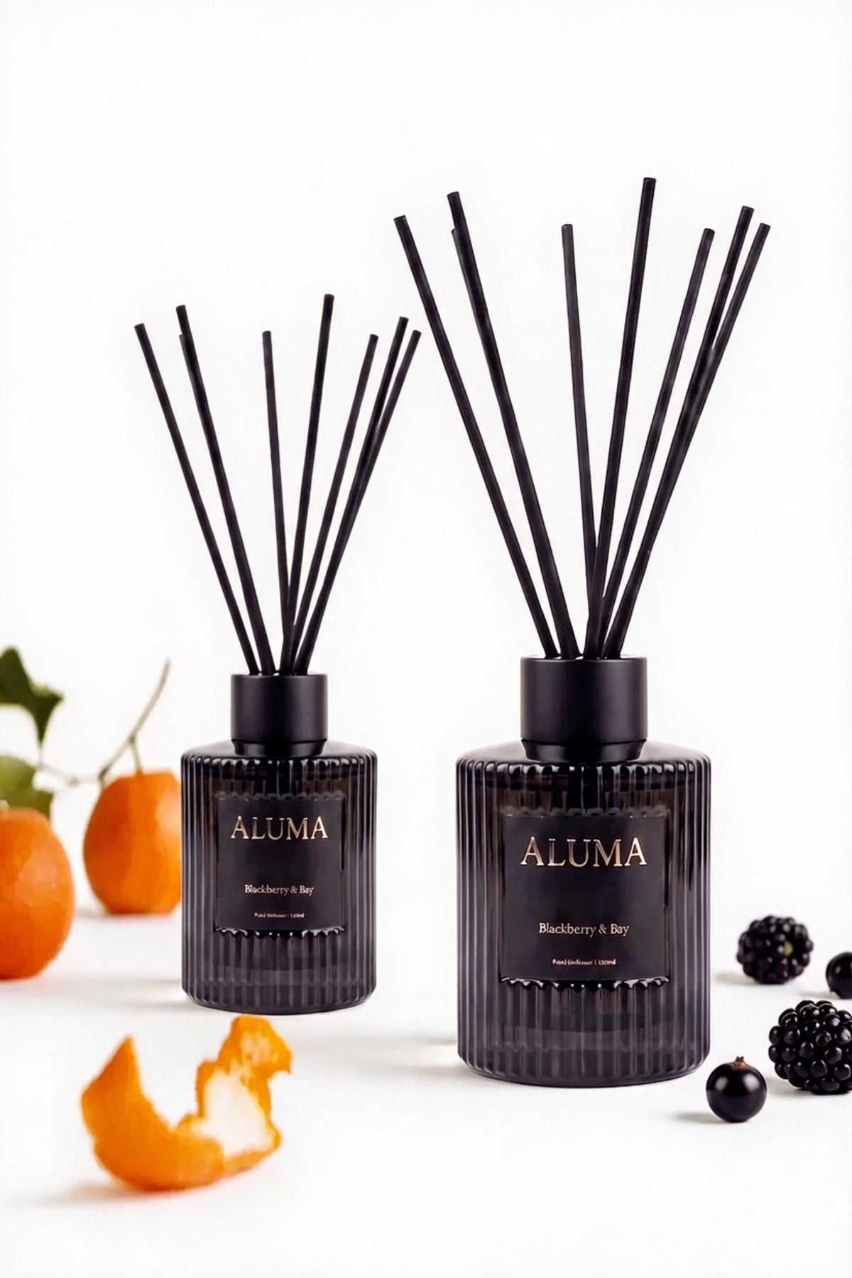 Two ALUMA reed diffusers with black glass bottles and black reeds, styled with citrus peels, oranges, and blackberries on a white background.