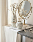 A styled interior scene with a round ornate gold-framed mirror on a dark table beside a draped white tablecloth. Two floral ceramic vases with dried white flowers sit on a marble tray, and a small matching floral pot is in front.