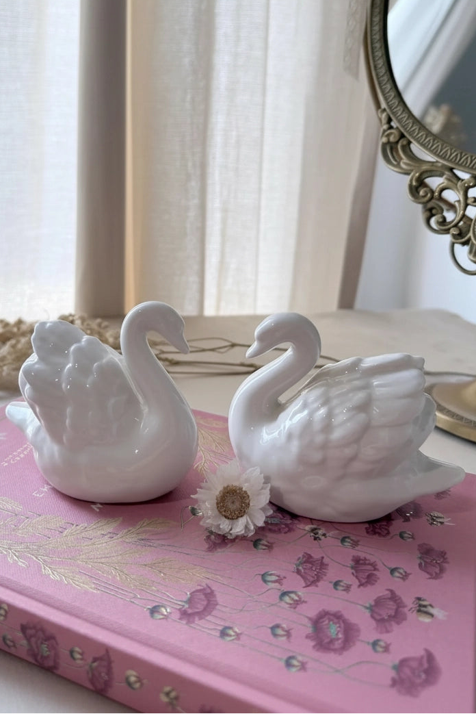 Two white ceramic swan figurines placed on a pink floral book, with a small white flower between them, in a softly lit indoor setting near a mirror.