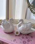 Two white ceramic swan figurines placed on a pink floral book, with a small white flower between them, in a softly lit indoor setting near a mirror.