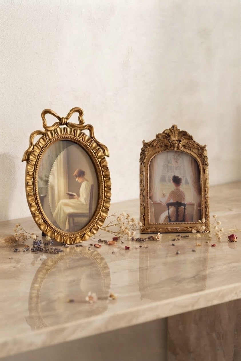 Two ornate gold-framed picture frames with vintage-style detailing, displayed on a light marble surface against a textured beige wall. The left frame is oval with a bow-like top ornament and contains a painting of a woman reading; the right frame is rectangular with a decorative crest top and contains a painting of a woman seated at a desk.