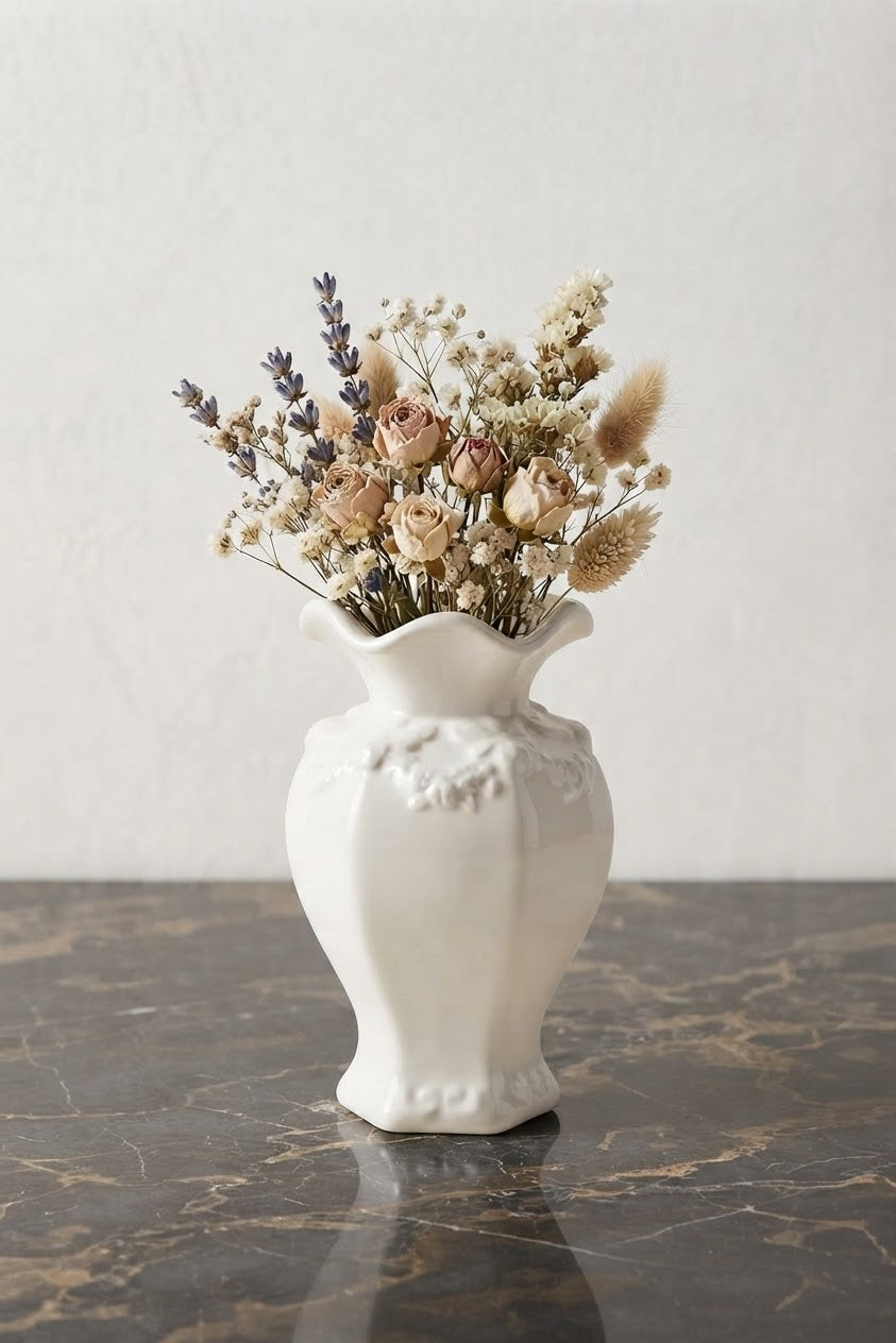 A white ceramic vase with a flared rim and embossed floral detailing, filled with dried flowers and grasses, set on a dark marble surface against a light textured wall.